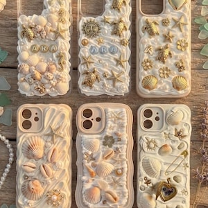 Puede incluir: Seis fundas para teléfono decoradas con una textura de crema blanca, con varios dijes dorados que incluyen conchas marinas, estrellas de mar, tortugas, un sol, un corazón y las letras A, R, A. Los diseños incorporan pequeñas perlas y detalles metálicos.