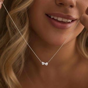 May include: A silver necklace with two heart-shaped pendants. The necklace is shown against a blurred background. The chain is fine and the hearts are smooth. The jewellery is simple and elegant.