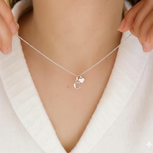 Puede incluir: Un collar plateado con una cadena fina. El collar presenta un pequeño dije con la letra "C" y un dije en forma de corazón. El collar se muestra sobre un fondo blanco.