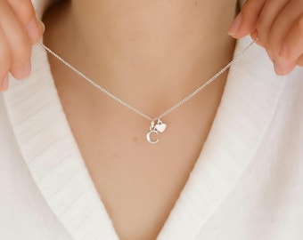 May include: A silver necklace with a fine chain. The necklace has a small charm with the letter "C" and a heart-shaped charm. The necklace is shown against a white background.