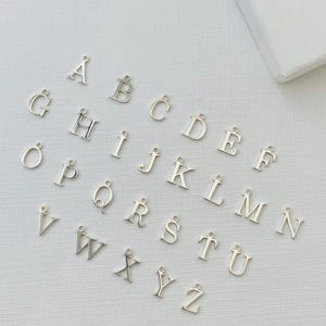 May include: Silver alphabet charms, each with a small loop for attaching to jewellery or other crafts. The letters are in a simple, sans-serif font and are scattered on a white surface. A white box is in the upper right corner.
