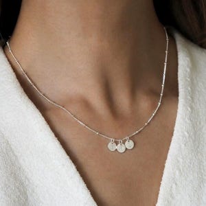 May include: A delicate silver necklace featuring three small, round pendants. Each pendant is engraved with a single letter. The necklace is worn around the neck, and the chain has a beaded design.