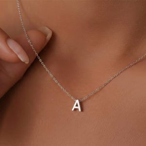 Puede incluir: Un delicado collar de plata con un colgante con la letra "A". El pequeño colgante cuelga de una fina cadena. El collar se muestra sobre un fondo de tono de piel, destacando el diseño minimalista de la joya.