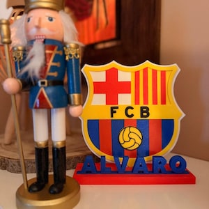 Può includere: Un soldato schiaccianoci in legno con uniforme blu e dettagli dorati è accanto a un cartello stampato in 3D. Il cartello mostra il logo del FC Barcelona e il nome "ALVARO" in lettere blu su una base rossa.