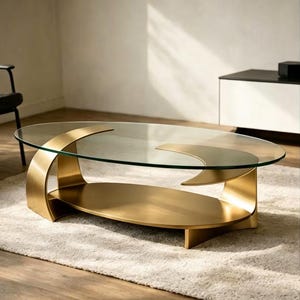 Puede incluir: Una mesa de centro ovalada con una tapa de cristal transparente y una base de metal dorado. La base tiene un diseño curvo único con un estante inferior adicional. La mesa mide aproximadamente 122 cm de largo y 61 cm de ancho.