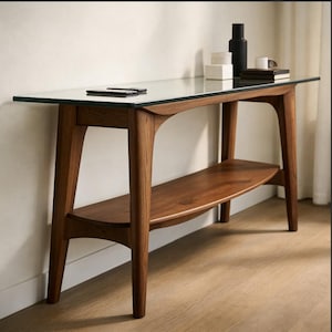 Op de afbeelding: Een console in mid-century modern stijl met een helder glazen blad en een onderste plank. De tafel is gemaakt van donker hout en heeft een strak, minimalistisch ontwerp. Een zwarte fles, boeken en een telefoon liggen op de tafel.