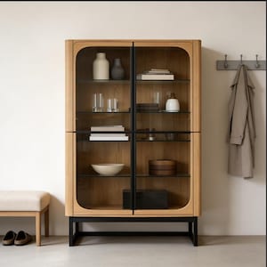 以下が含まれることがあります： ガラス扉と黒い金属製の脚が付いた木製のディスプレイキャビネット。キャビネットには、花瓶、本、ボウルなどの装飾品が詰められています。背景にはベンチとコートラックがあり、リビングルームまたは玄関を示唆しています。