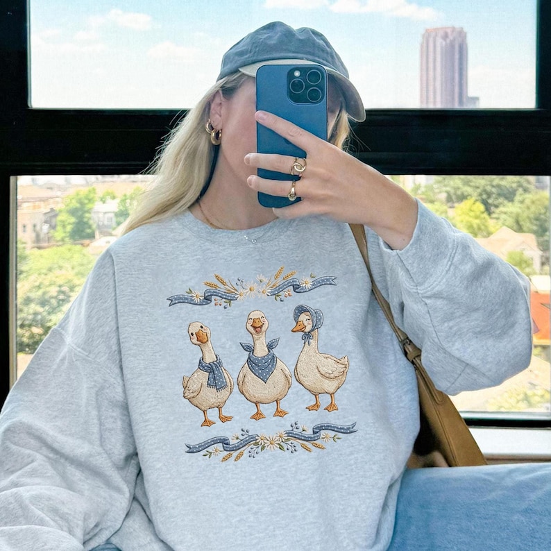 May include: Light gray sweatshirt featuring a whimsical illustration of three geese. The geese are adorned with blue scarves and bonnets, set against a backdrop of wheat and floral accents. The sweatshirt is a comfortable, casual style.