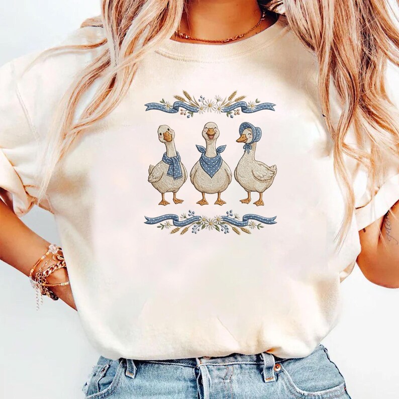 May include: Cream-colored t-shirt featuring a whimsical illustration of three geese. Each goose is adorned with a blue scarf or bonnet. The design is framed by blue ribbons and floral accents, creating a charming, pastoral aesthetic.
