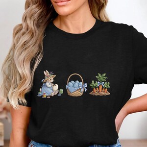 May include: Black t-shirt featuring an Easter-themed graphic. The design includes a bunny in a blue dress, a basket of blue eggs, and carrots. The bunny is wearing a hat and painting an egg. The basket has a blue bow.