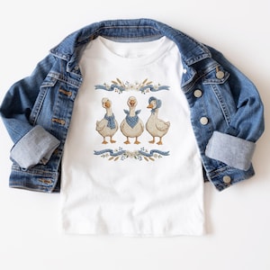 May include: White t-shirt featuring a whimsical illustration of three geese adorned with blue bandanas and a bonnet. The geese are set against a backdrop of floral and ribbon elements. A denim jacket is draped over the shirt.