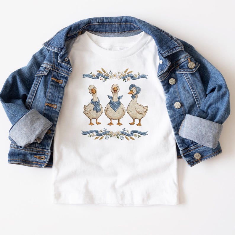 May include: White t-shirt featuring a whimsical illustration of three geese adorned with blue bandanas and a bonnet. The geese are set against a backdrop of floral and ribbon elements. A denim jacket is draped over the shirt.
