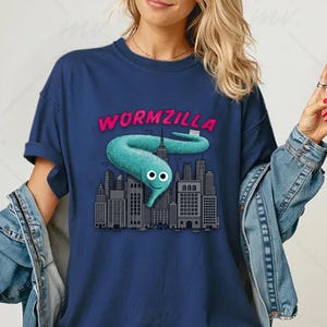 Puede incluir: Camiseta azul marino con un personaje de gusano turquesa y el texto rosa "WORMZILLA" sobre un paisaje urbano. El gusano tiene ojos saltones y una cara sonriente. La camiseta tiene las mangas enrolladas.