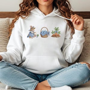 May include: White hoodie featuring an Easter-themed graphic. The design includes a bunny, a basket of blue eggs, and carrots. The bunny is wearing a blue dress and hat. The graphic is in soft pastel colors.