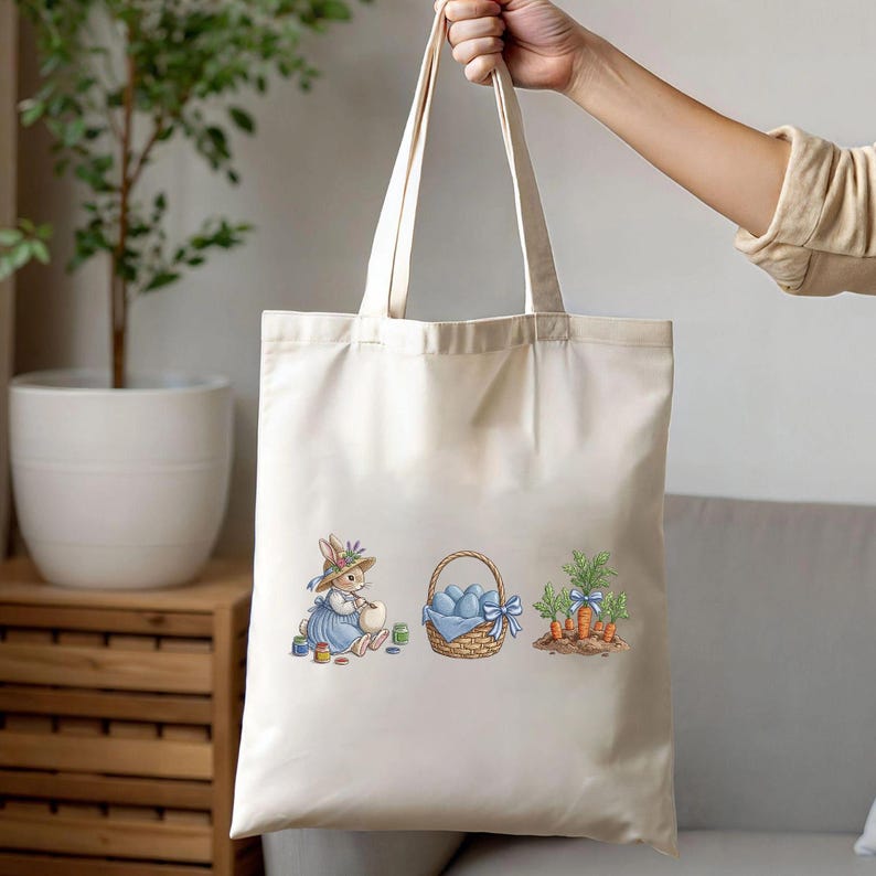 May include: Cream-colored tote bag featuring an Easter theme. The design includes a bunny painting eggs, a basket of blue eggs, and carrots with blue bows. The bag has two handles and is held up by a person.