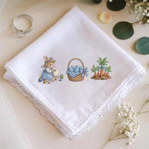 May include: White handkerchief with a delicate lace edge. The handkerchief features an embroidered Easter scene with a bunny painting eggs, a basket of blue eggs, and carrots. A silver ring is in the upper left corner.