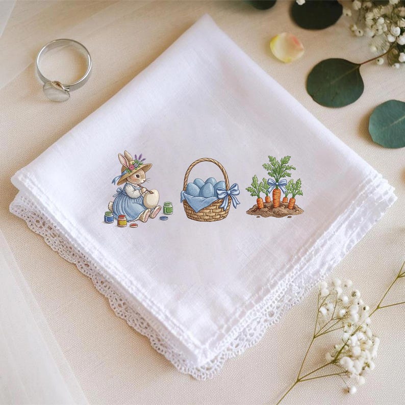 May include: White handkerchief with a delicate lace edge. The handkerchief features an embroidered Easter scene with a bunny painting eggs, a basket of blue eggs, and carrots. A silver ring is in the upper left corner.