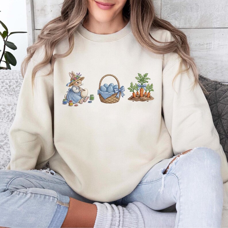 May include: Cream-colored sweatshirt featuring an Easter-themed graphic. The design includes a bunny in a blue dress, a basket of blue eggs tied with a blue bow, and a patch of carrots with green tops. The sweatshirt is a comfortable, casual style.