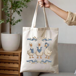 May include: Cream-colored tote bag featuring an embroidered design of three geese wearing blue scarves and a bonnet. The geese are positioned below blue ribbons and floral accents. The bag is held up by a person.