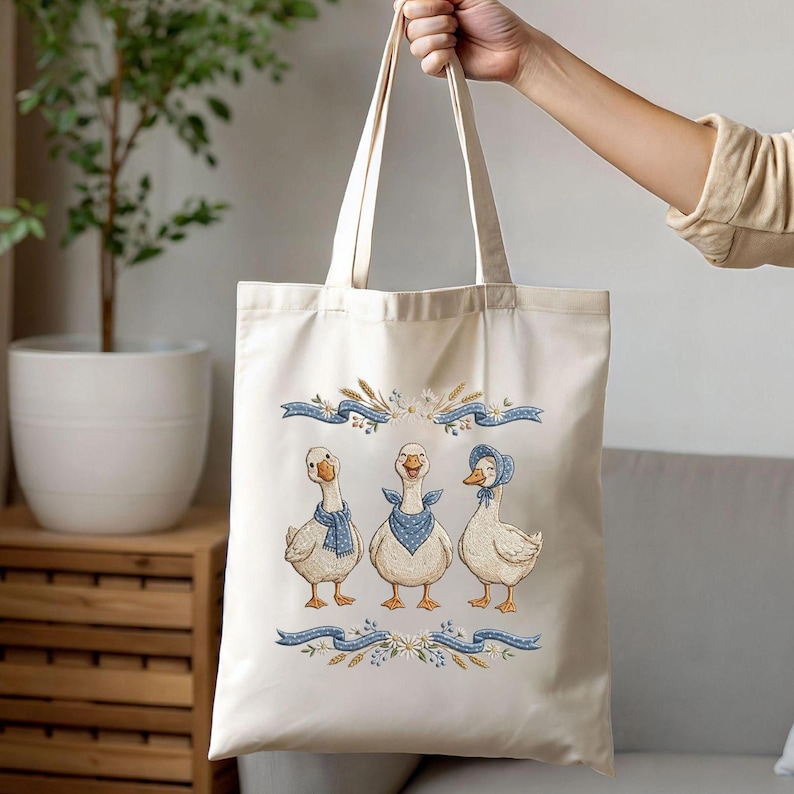 May include: Cream-colored tote bag featuring an embroidered design of three geese wearing blue scarves and a bonnet. The geese are positioned below blue ribbons and floral accents. The bag is held up by a person.