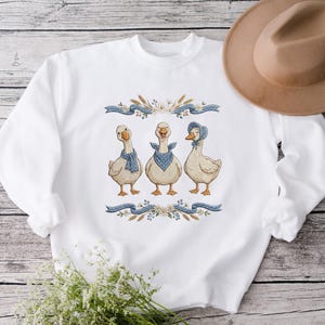 May include: White sweatshirt featuring an embroidered design of three geese wearing blue scarves and bonnets. The geese are framed by blue ribbons, wheat, and floral accents. A tan hat rests on the side.