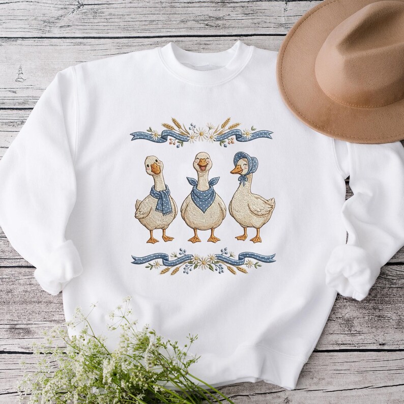 May include: White sweatshirt featuring an embroidered design of three geese wearing blue scarves and bonnets. The geese are framed by blue ribbons, wheat, and floral accents. A tan hat rests on the side.