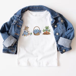 May include: White t-shirt featuring an Easter-themed design. The design includes a bunny in a blue dress, a basket of blue eggs, and carrots. A denim jacket is draped over the shirt.