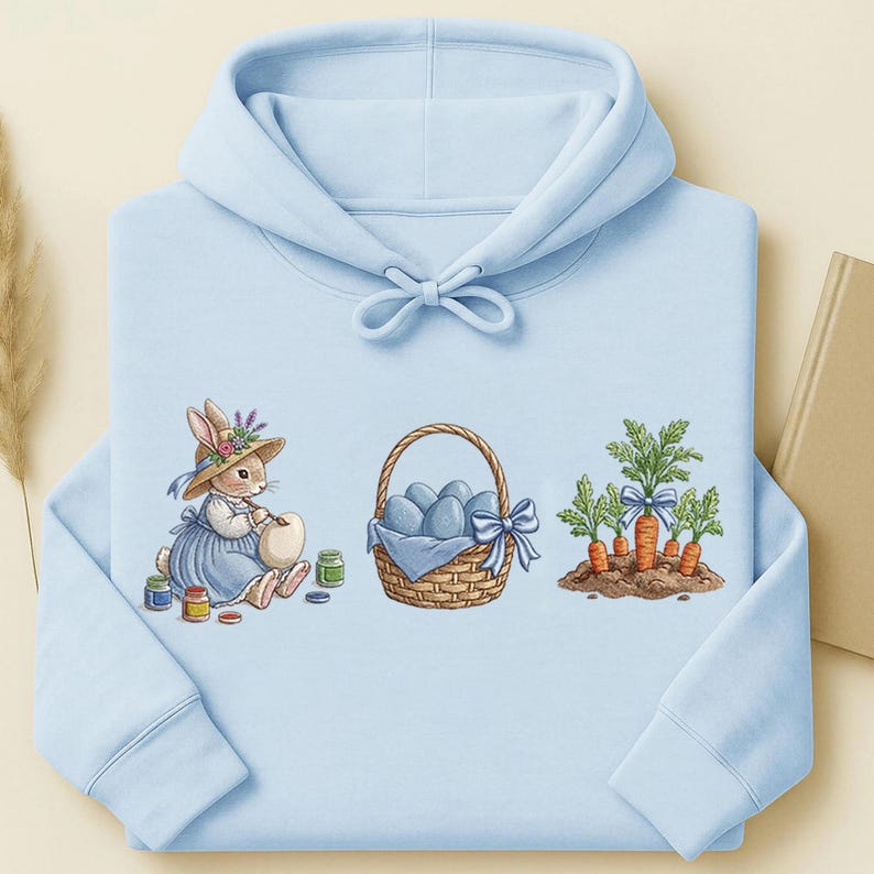 May include: Light blue hoodie with an Easter-themed design. The design features a bunny painting an egg, a basket of blue eggs, and carrots with green tops. The hoodie has a drawstring hood and long sleeves.