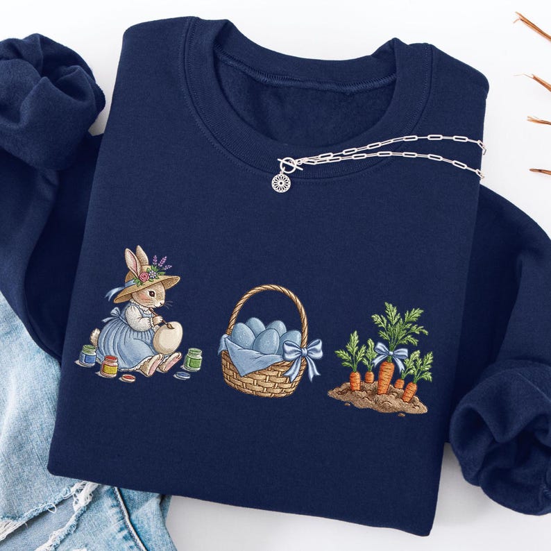 May include: Navy blue sweatshirt with an embroidered Easter design. The design features a bunny painting an egg, a basket of blue eggs, and a patch of carrots. A silver necklace with a sun pendant is draped across the sweatshirt.