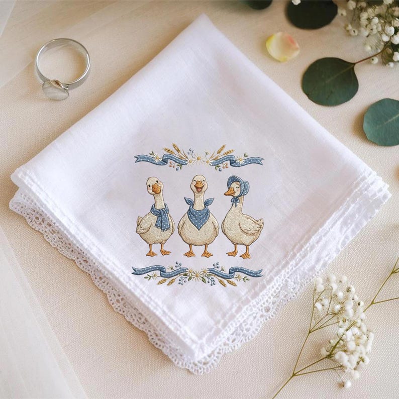 May include: White handkerchief with a delicate lace edge, featuring an embroidered design of three geese wearing blue scarves and bonnets. The embroidery includes blue ribbons and wheat stalks. A silver ring is in the upper left corner.