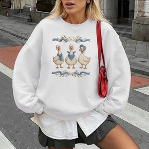 May include: White sweatshirt featuring a whimsical illustration of three geese wearing blue scarves and a bonnet. The geese are centered on the front, surrounded by blue ribbons and floral accents. The sweatshirt has long sleeves and a crew neck.