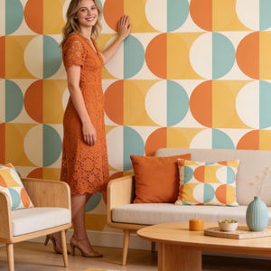 Peut inclure: Une femme en robe en dentelle orange se tient devant un mur avec un motif géométrique rétro orange, jaune et turquoise. La pièce comprend un canapé, des chaises et une table basse, le tout dans un style moderne du milieu du siècle. Un tapis coloré complète le design mural.