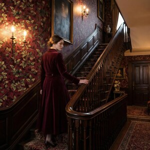 Peut inclure: Une personne v&ecirc;tue d'un manteau de velours bordeaux descend un escalier en bois dans une maison d'&eacute;poque. Les murs sont orn&eacute;s de papier peint floral et d'&oelig;uvres encadr&eacute;es. L'escalier a une rampe et des marches en bois fonc&eacute;.