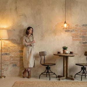 Op de afbeelding: Een vrouw in een beige jurk staat bij een bakstenen muur in een kamer met een ronde houten tafel en twee stoelen. Een vloerlamp en een hanglamp verlichten de ruimte. Potplanten en een geweven vloerkleed dragen bij aan de inrichting.