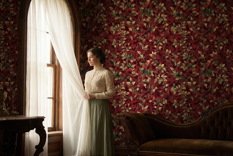Peut inclure: Une femme v&ecirc;tue d'une robe vintage se tient pr&egrave;s d'une fen&ecirc;tre avec des rideaux blancs. La pi&egrave;ce pr&eacute;sente un papier peint floral rouge et une table en bois sombre. La robe est de couleur claire avec un col montant et des manches longues.