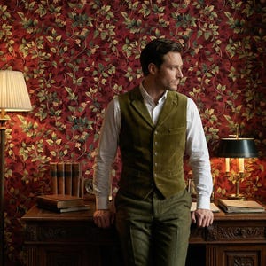 Peut inclure: Un homme porte un gilet vert et un pantalon assorti dans une pi&egrave;ce avec un papier peint floral rouge. Un bureau avec des livres et une lampe sont devant lui. Des biblioth&egrave;ques sont de chaque c&ocirc;t&eacute; de la pi&egrave;ce.