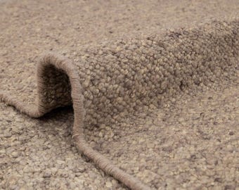 May include: Close-up of a rolled-up, textured rug in shades of beige and brown. The rug's surface has a nubby, speckled appearance, with a visible border. The rolled edge reveals the rug's thickness and construction. Suitable for home decor.