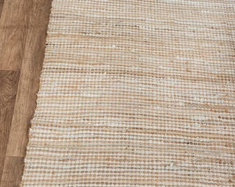 Peut inclure: Tapis rectangulaire en jute tissé, aux tons naturels. Il présente un motif rayé texturé et une frange sur un bord. Le tapis est posé sur un sol en bois clair.