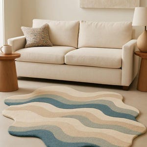 Op de afbeelding: Een golvend, abstract tapijt in blauw, beige en bruintinten. Het tapijt ligt op een lichte vloer voor een crèmekleurige bank. Het ontwerp lijkt op oceaangolven en voegt een moderne touch toe aan de kamer. Afmetingen ca. 180 cm x 120 cm.