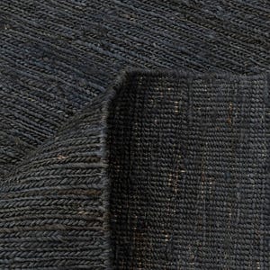 May include: Close-up of a dark grey woven rug with a textured surface. The rug is made of thick, braided fibres, with a folded corner revealing the underside. The weave pattern creates diagonal lines across the surface, giving it a rustic appearance.