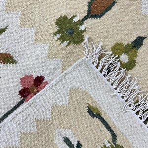 Op de afbeelding: Close-up van een beige en wit patroonkleed met bloemen- en geometrische ontwerpen. Het kleed heeft groene, bruine en roze bloemmotieven. Het kleed heeft een gefranjerde rand en een getextureerd oppervlak.