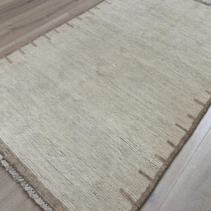 Könnte beinhalten: Ein rechteckiger beigefarbener Teppich mit strukturierter Oberfläche und braunem Rand. Der Teppich hat ein schlichtes Design mit vertikalen Linien und Fransen an einem Ende. Der Rand hat gleichmäßig verteilte rechteckige Akzente.