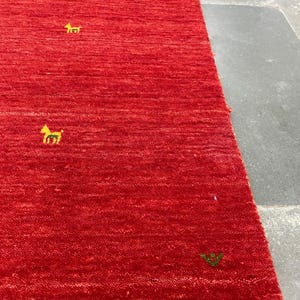 Puede incluir: Una alfombra roja vibrante con ribetes amarillos y pequeños motivos bordados de animales y plantas. La alfombra tiene una superficie texturizada y una forma rectangular. Las dimensiones de la alfombra no son visibles en la imagen.