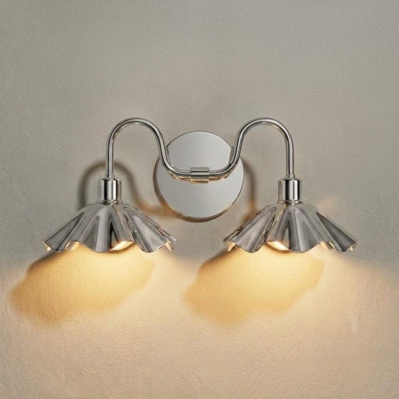 Polished chrome double wall sconce illuminated with warm bulbs, casting golden light beneath two pleated metal shades onto a textured neutral wall.