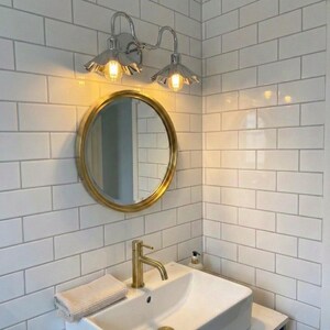 Chrome double wall sconce mounted above a round gold-framed mirror on white subway tiles in a bathroom, with two warm glowing bulbs and a white sink below.