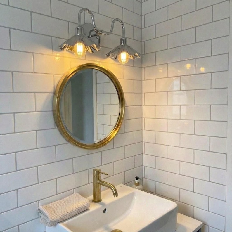 Chrome double wall sconce mounted above a round gold-framed mirror on white subway tiles in a bathroom, with two warm glowing bulbs and a white sink below.