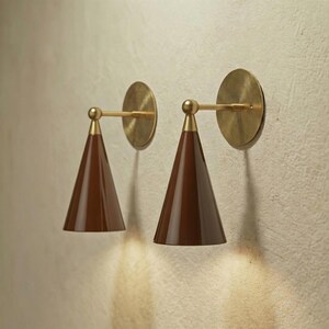 Peut inclure: Deux appliques murales de style moderne. Chaque applique est composée d'un abat-jour conique marron, d'un bras en laiton et d'une plaque murale circulaire en laiton. Elles sont fixées sur un mur texturé blanc cassé.
