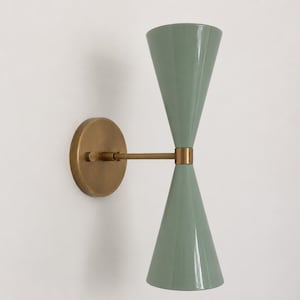 Puede incluir: Un aplique de pared con dos pantallas cónicas de color verde salvia dispuestas verticalmente. Las pantallas están conectadas por un brazo horizontal central de tono latón que se fija a una placa de pared circular de latón con un pequeño pomo.