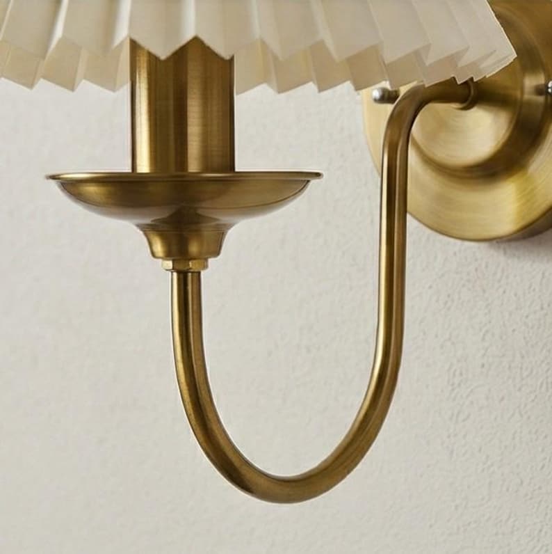 May include: A wall-mounted sconce with a brass finish. The fixture features a pleated, off-white lampshade and a curved arm. The design is classic and elegant, suitable for various interior styles. The sconce is mounted on a white wall.