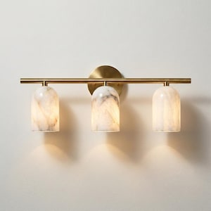 Puede incluir: Un aplique de pared de tres luces con una barra horizontal de latón cepillado y un soporte circular. Tres pantallas en forma de cúpula hechas de piedra veteada blanca y gris cuelgan de la barra, emitiendo un brillo suave y cálido hacia abajo.